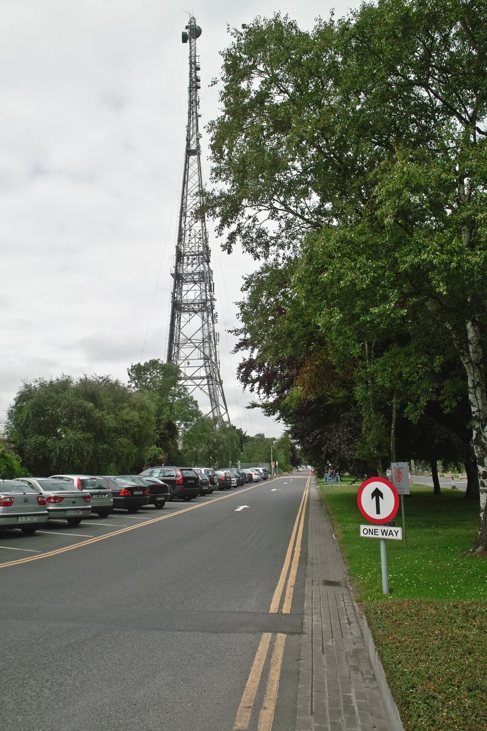A broadcasting mast beside a road lined with parked cars, with trees on either side.
