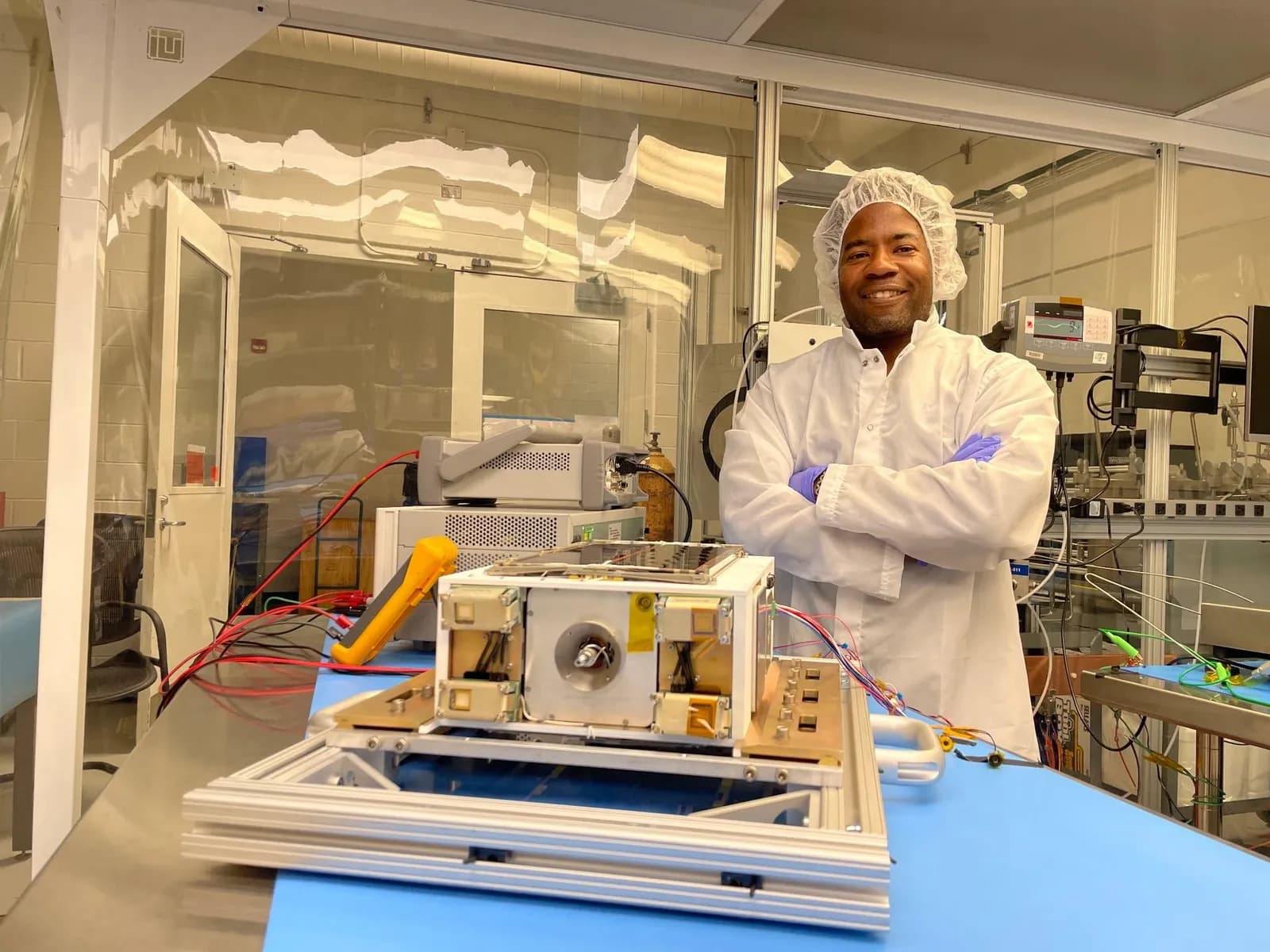 Nehemiah Williams stands behind CubeSat flight hardware and test equipment in a clean room, wearing a white lab coat, hair covering and blue gloves.