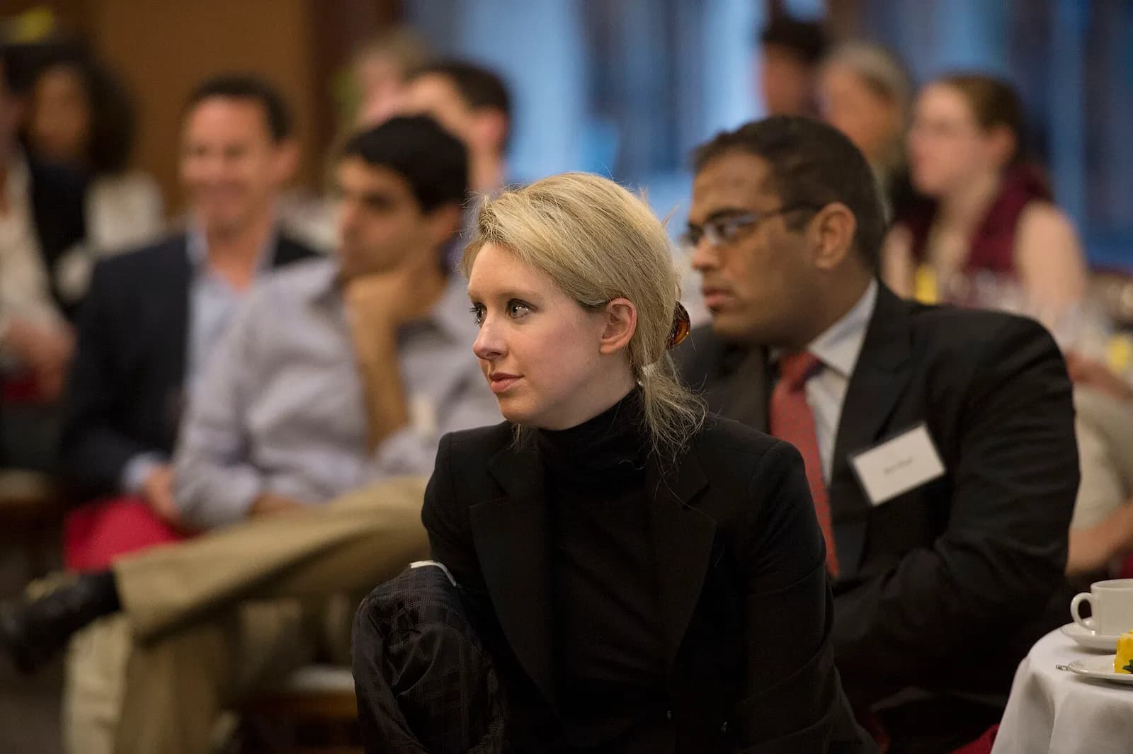 A blonde woman in a black jacket and black turtleneck looking to her left while seated in an audience, with blurred attendees behind her.