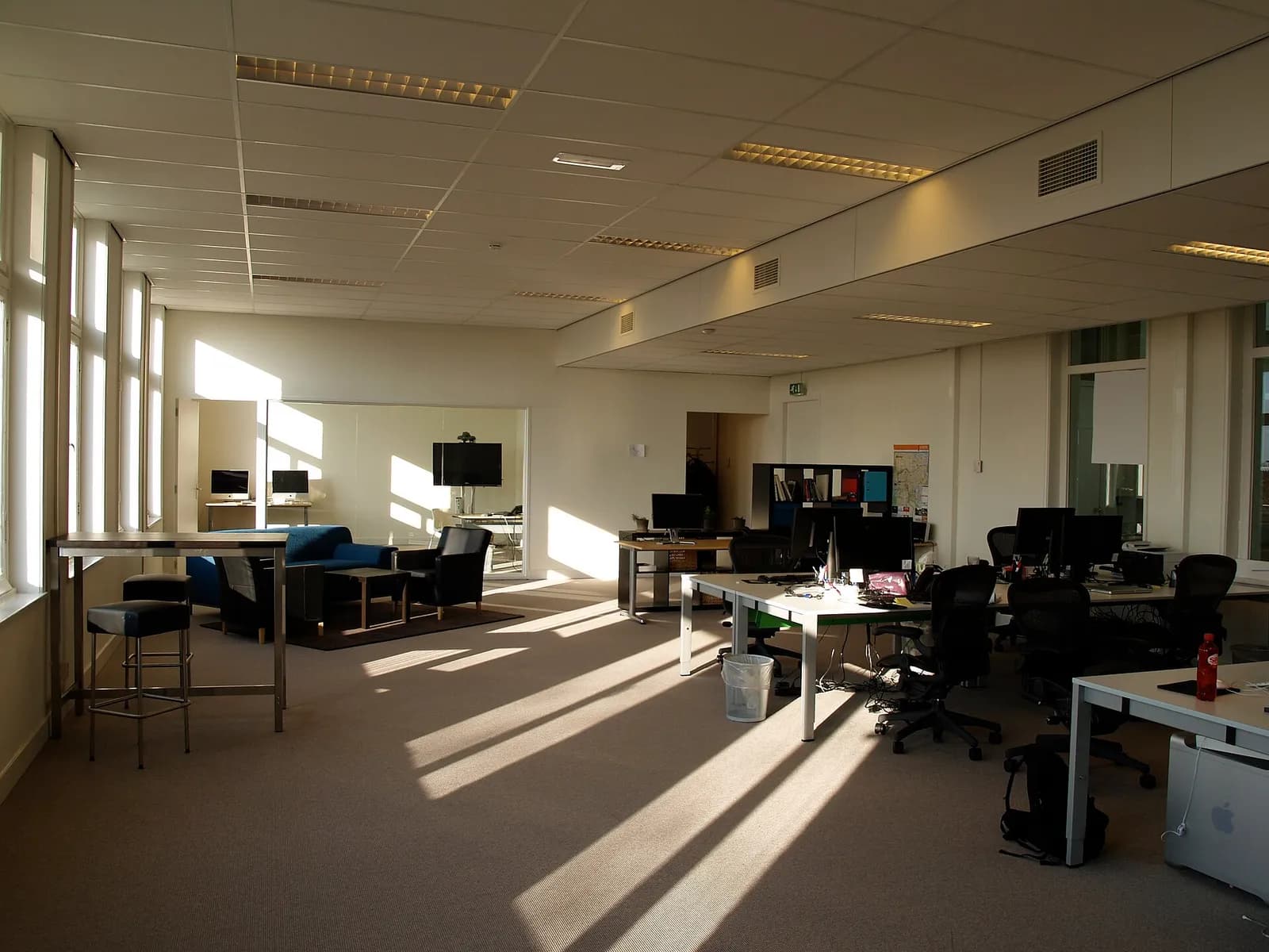 Office interior with desks, chairs and computer monitors beside tall windows.